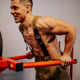 Uśmiechnięty mężczyzna w stroju strażackim ćwiczy pompki na poręczach, eksponując wyrzeźbioną sylwetkę i tatuaż na boku.