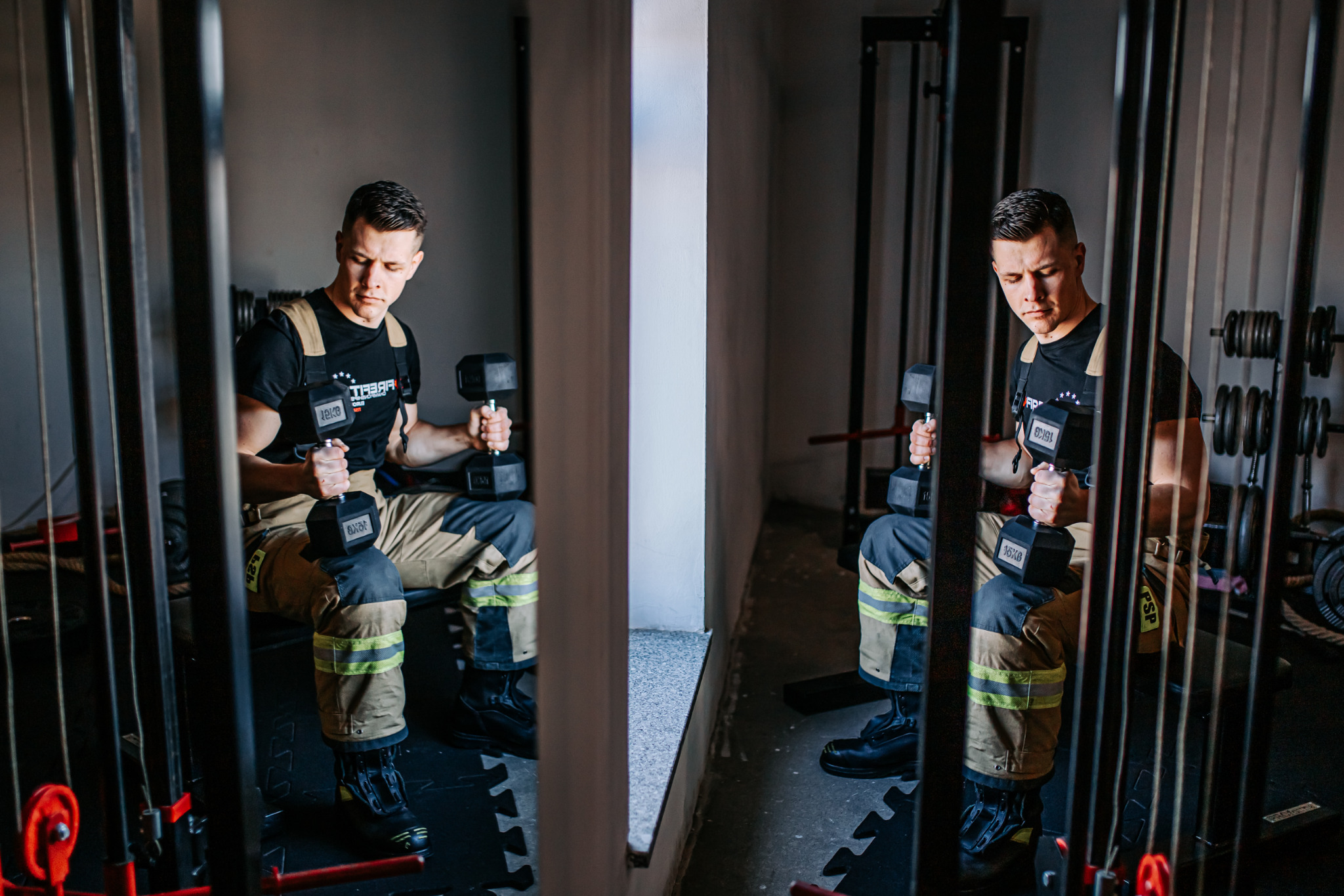 Mężczyzna w stroju strażackim siedzi na ławce i podnosi dwie czarne hantle 16kg w siłowni, widoczne elementy wyposażenia siłowni.