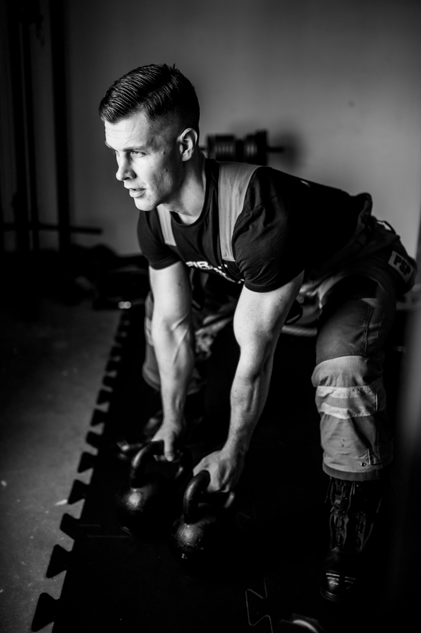 Czarno-białe zdjęcie mężczyzny w stroju strażackim, pochylonego nad dwoma odważnikami kettlebell na podłodze w siłowni w Świdnicy, przygotowującego się do podniesienia ciężarów.