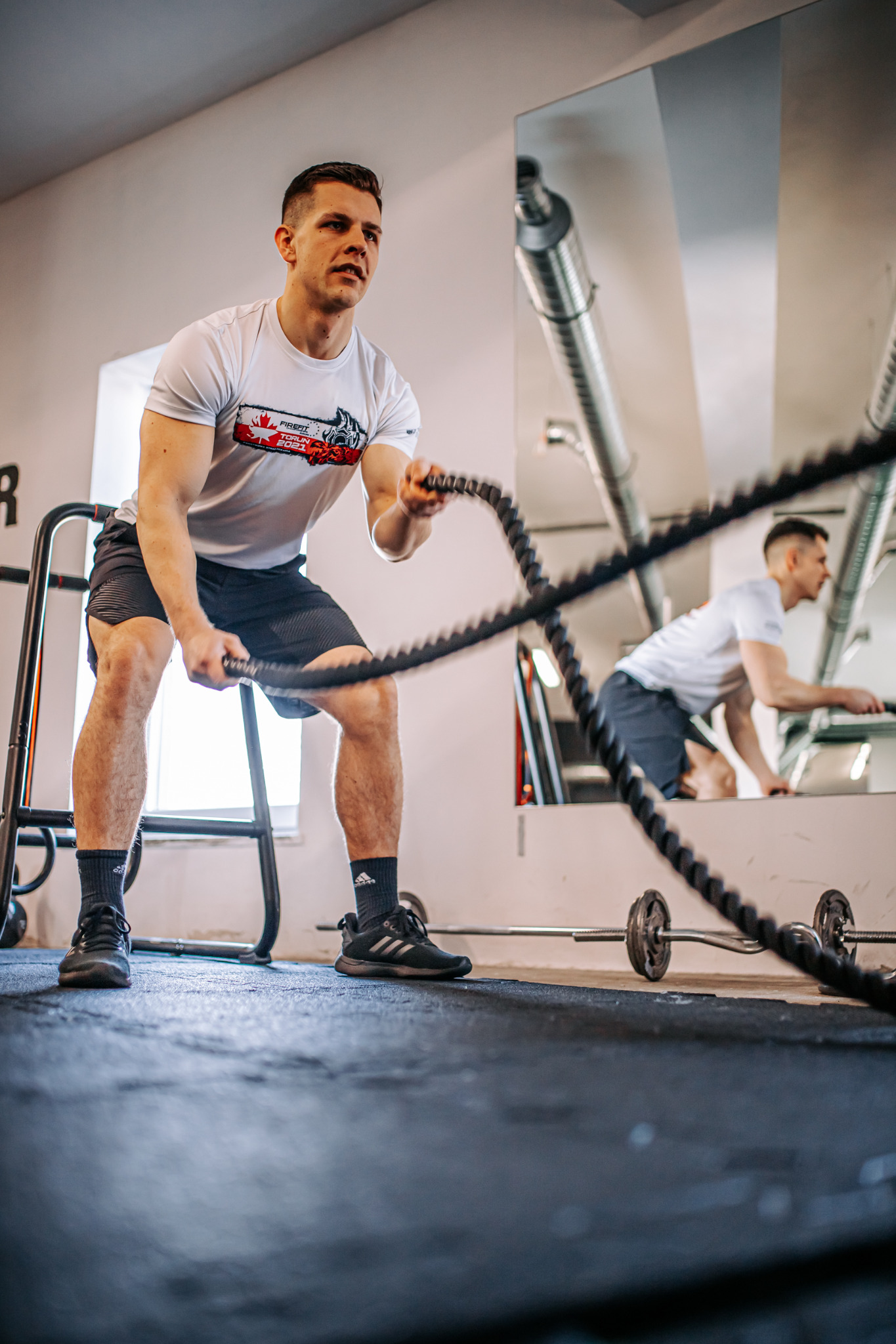 Mężczyzna w sportowej koszulce z krótkim rękawem ćwiczy z grubymi linami w siłowni, odbicie w lustrze pokazuje go w trakcie ćwiczenia z hantlami.
