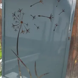 Szara metalowa brama wjazdowa z wyciętym wzorem dmuchawca, widoczne zawiasy i drewniane ogrodzenie w tle.