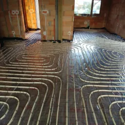 Rozłożona instalacja ogrzewania podłogowego w surowym stanie, rury ułożone w pętle na folii z siatką, widoczne ściany z cegły dziurawki i okno.