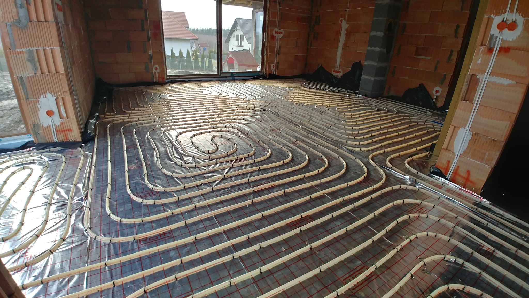 Widok z góry na rozłożone rury ogrzewania podłogowego w surowym wnętrzu, z widoczną folią i siatką montażową, oraz fragmentem ściany z cegły ceramicznej dziurawki.