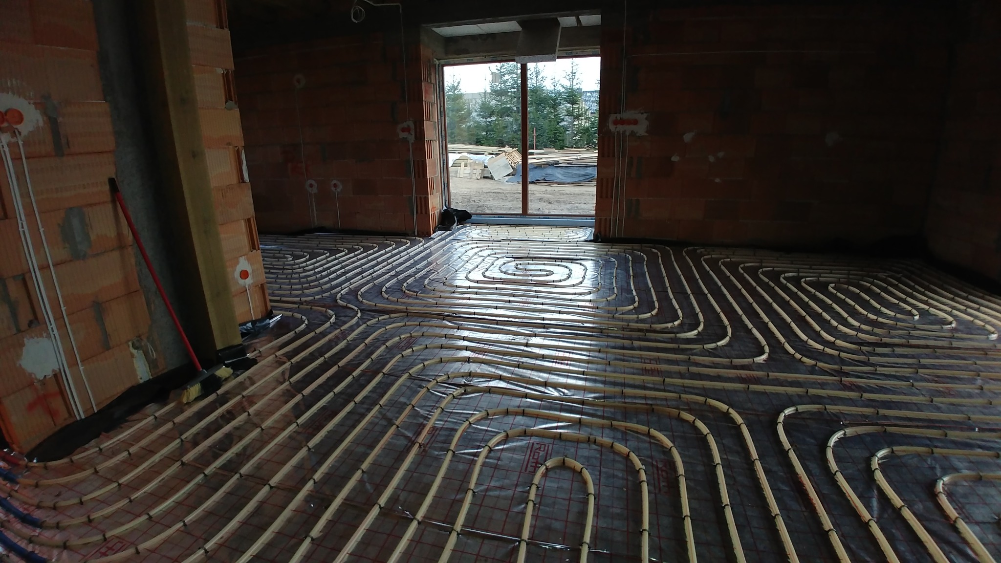 Rozłożona instalacja ogrzewania podłogowego z rurkami mocowanymi do siatki, widok na surowy stan budynku z pustakami i oknem na zewnątrz.