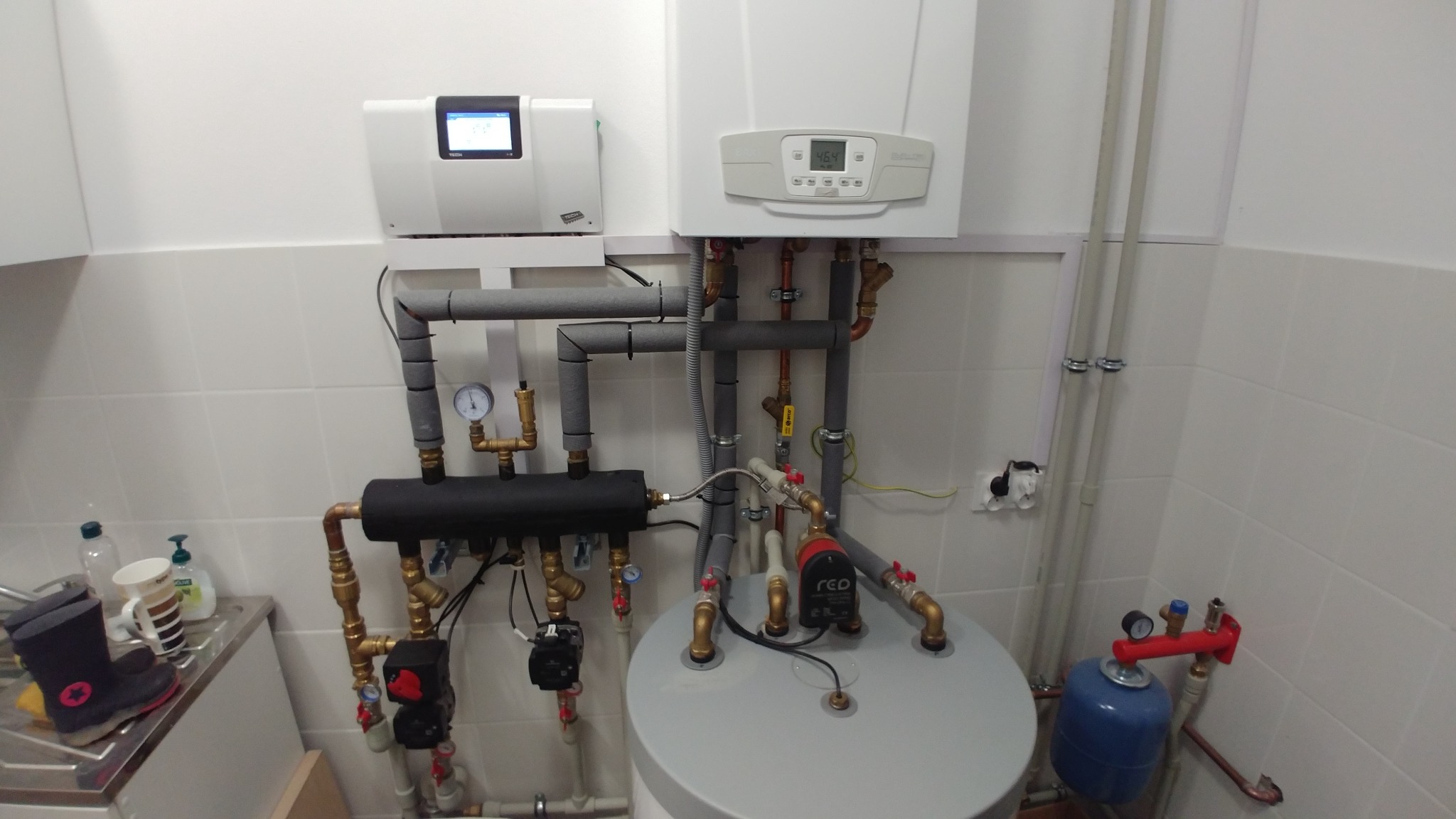 Kompletna instalacja grzewcza z piecem gazowym, zbiornikiem akumulacyjnym i rozdzielaczem, widoczna na tle białych płytek w pomieszczeniu technicznym. Dodatkowo widoczny biały panel sterowania...