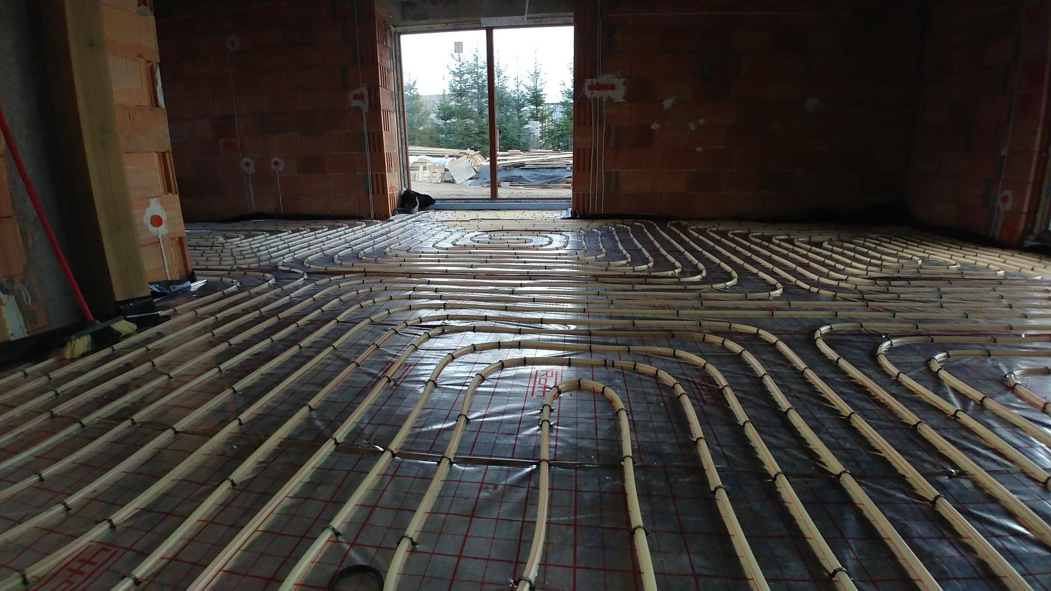 Instalacja ogrzewania podłogowego w surowym stanie, rury grzewcze ułożone na folii z siatką, widoczne drewniane listwy mocujące i niewykończone ściany z cegły.