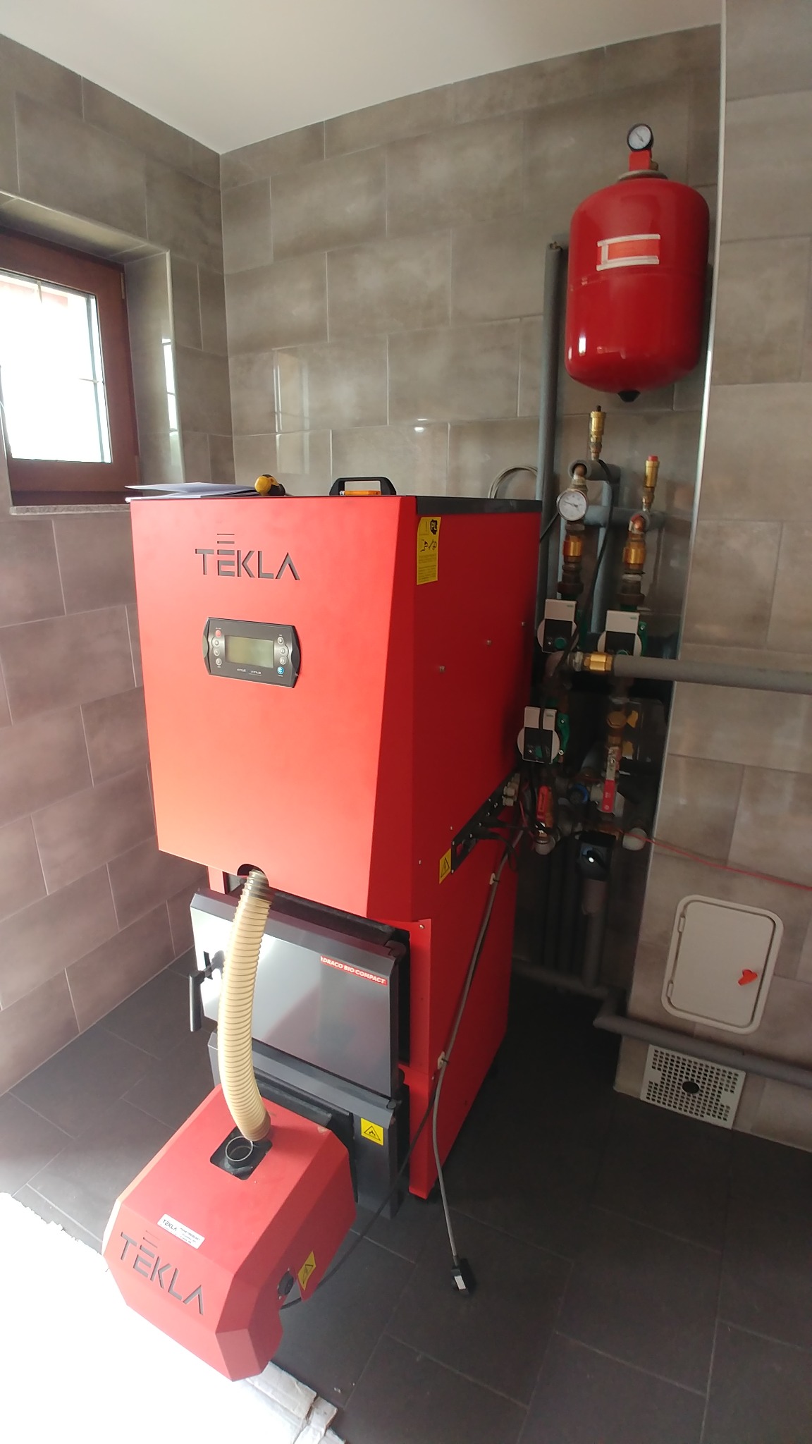 Czerwony piec na paliwo stałe marki Tekla, zainstalowany w pomieszczeniu z szarymi płytkami, widoczny zbiornik wyrównawczy i fragment instalacji.