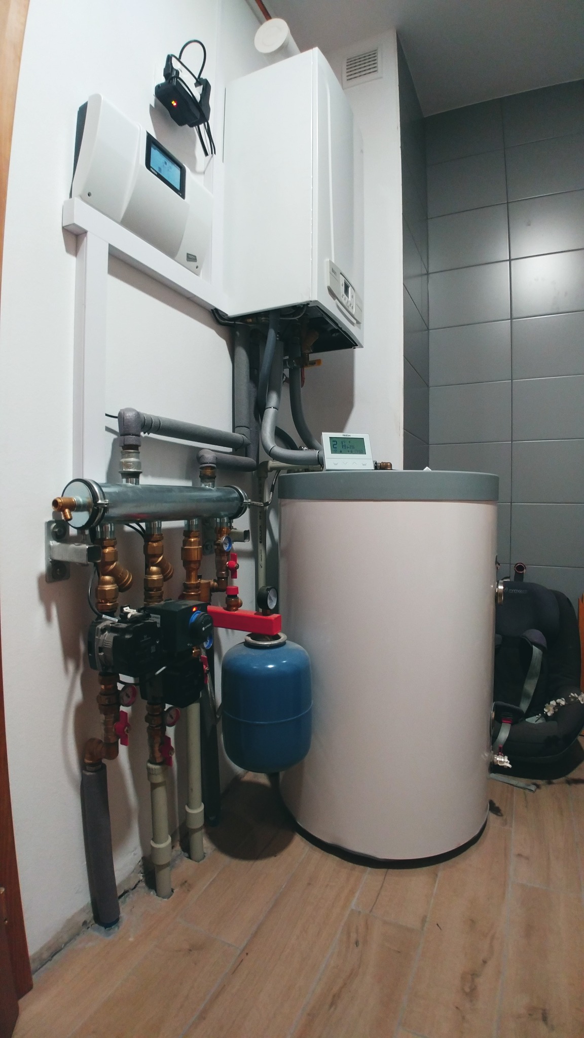 Zainstalowany, nowoczesny piec gazowy z białym zasobnikiem ciepłej wody, widoczne rury, zawory, manometry, sterownik i czujnik temperatury w łazience wyłożonej szarymi płytkami, na podłodze...