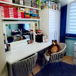 Gabinet z biurkiem, krzesłami obitymi szarą tkaniną, pluszowym misiem siedzącym na jednym z krzeseł oraz półkami z książkami i pomocami edukacyjnymi, w tym model szczęki i książka o ćwiczeniach...