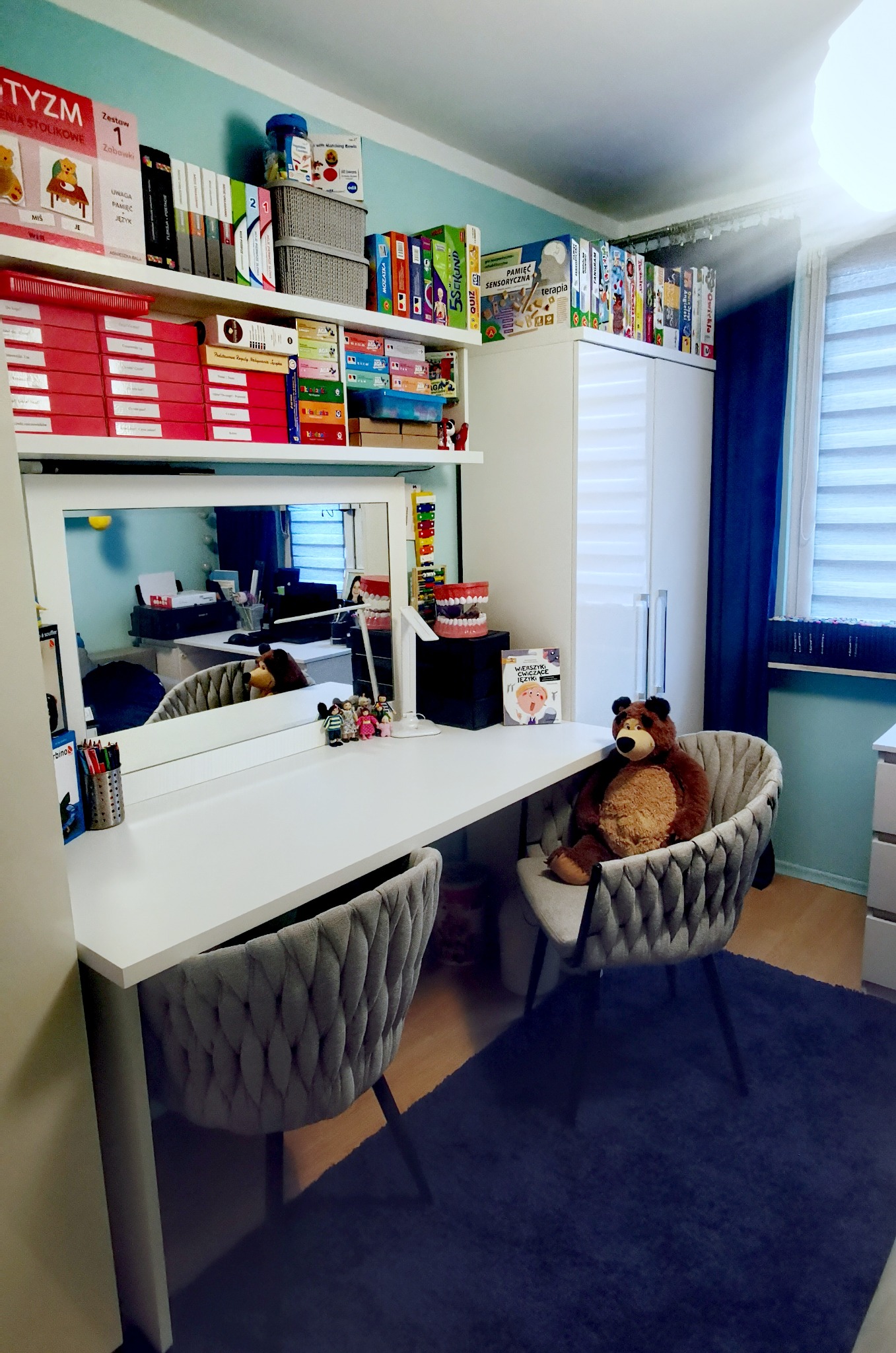Gabinet z biurkiem, krzesłami obitymi szarą tkaniną, pluszowym misiem siedzącym na jednym z krzeseł oraz półkami z książkami i pomocami edukacyjnymi, w tym model szczęki i książka o ćwiczeniach...