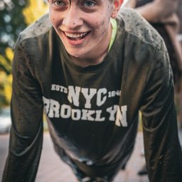 pietruszka tworzy - Uśmiechnięty nastolatek w trakcie wykonywania pompek na mokrym chodniku, ubrudzony ziemią, z kobietą w tle. Dynamiczne ujęcie z dołu.