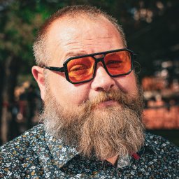 pietruszka tworzy - Portret brodatego mężczyzny w pomarańczowych okularach, ubranego w kwiecistą koszulę, na tle rozmytej zieleni. Nietypowe ujęcie, wyraziste kolory.