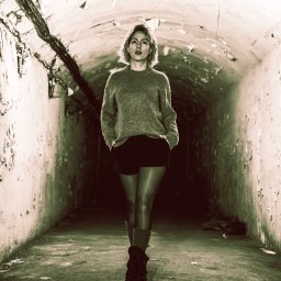 pietruszka tworzy - Kobieta w swetrze i szortach pozuje w tunelu z odpadającą farbą, oświetlonym od góry. Ujęcie centralne, podkreślające surowość otoczenia i kontrast z postacią.