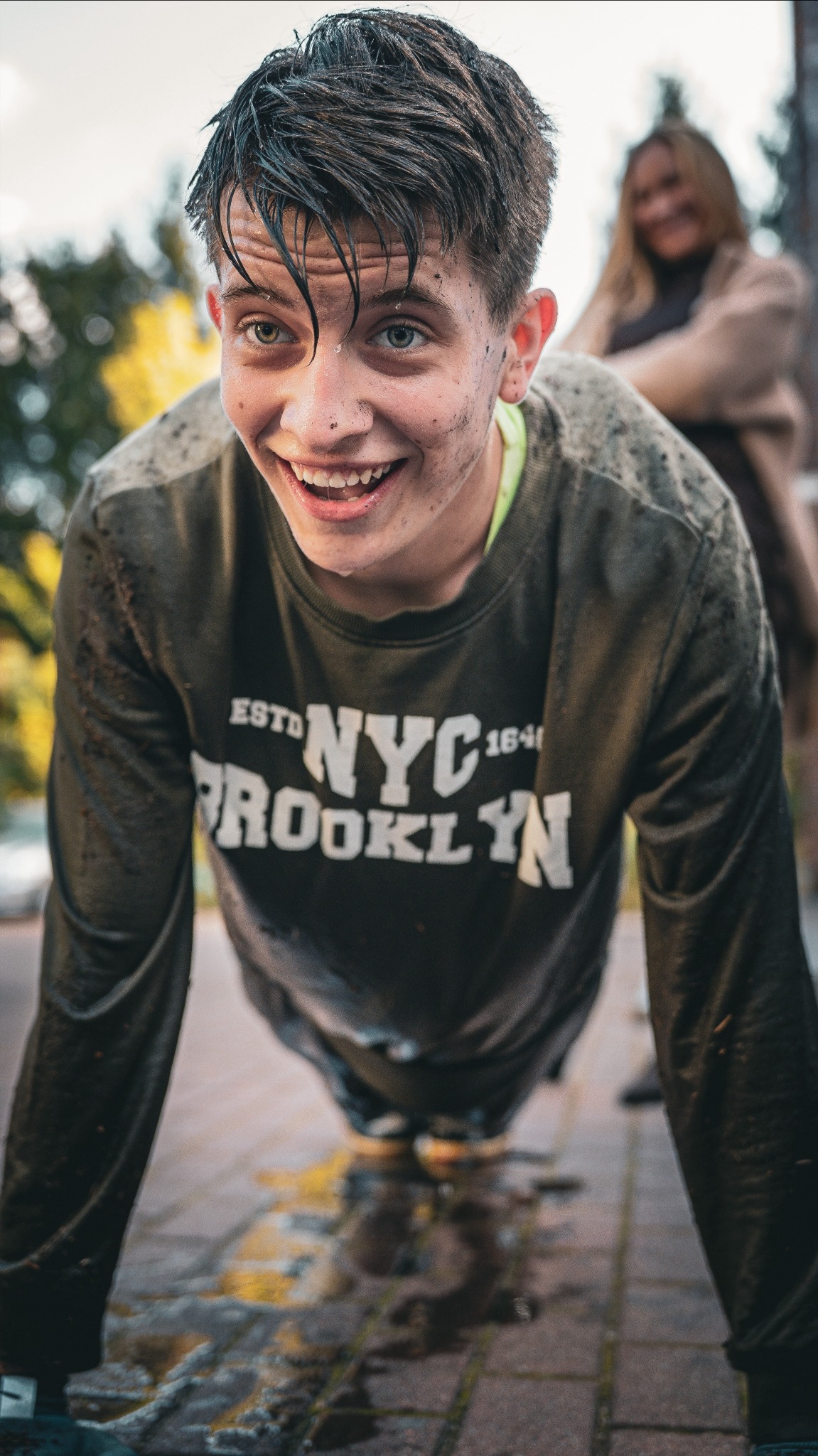 Uśmiechnięty nastolatek w trakcie wykonywania pompek na mokrym chodniku, ubrudzony ziemią, z kobietą w tle. Dynamiczne ujęcie z dołu.