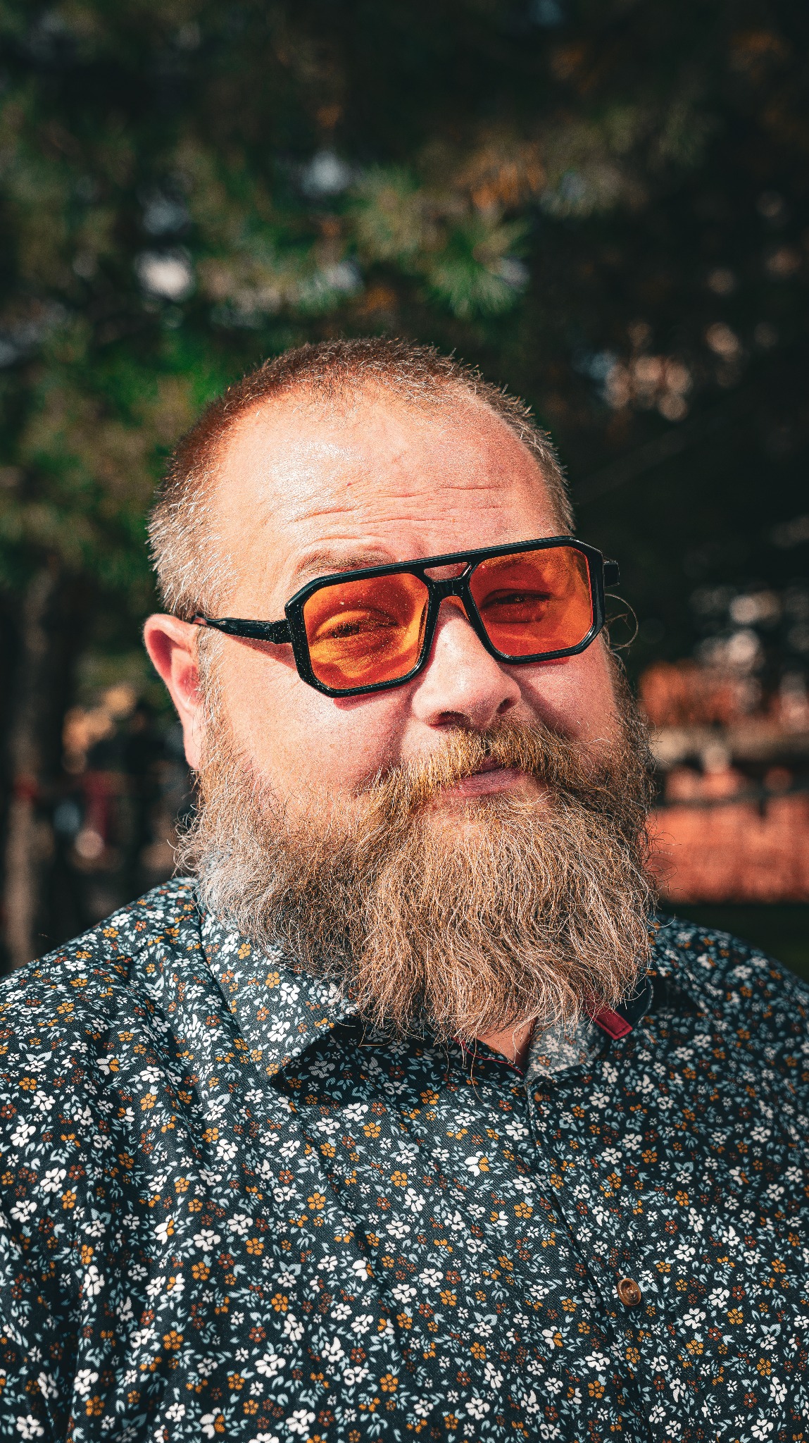 Portret brodatego mężczyzny w pomarańczowych okularach, ubranego w kwiecistą koszulę, na tle rozmytej zieleni. Nietypowe ujęcie, wyraziste kolory.
