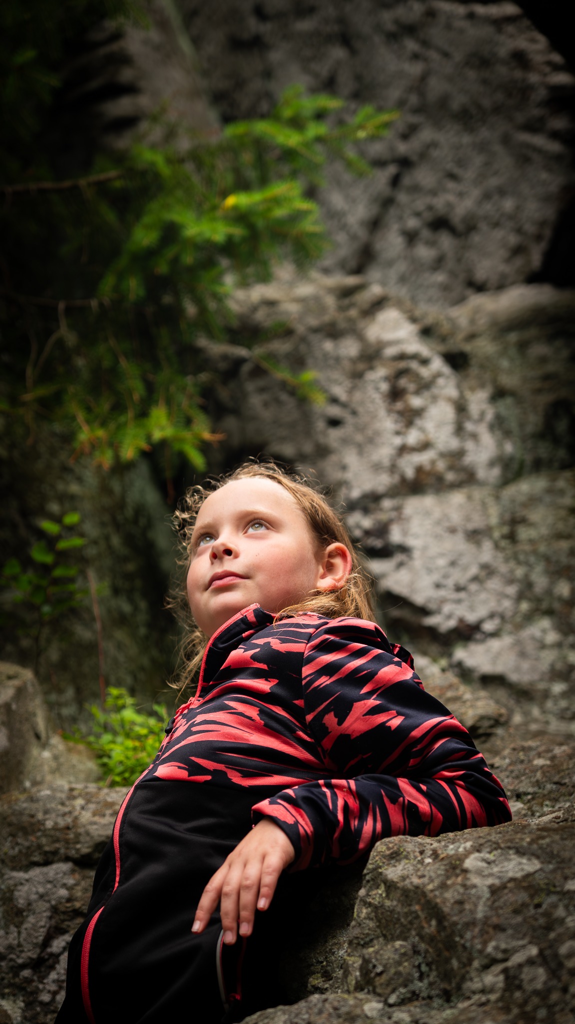 Portret dziewczynki w czarno-czerwonej bluzie, spoglądającej w górę na tle skalnej ściany z zielonymi akcentami roślinności. Ujęcie z dołu, podkreślające naturalne otoczenie.