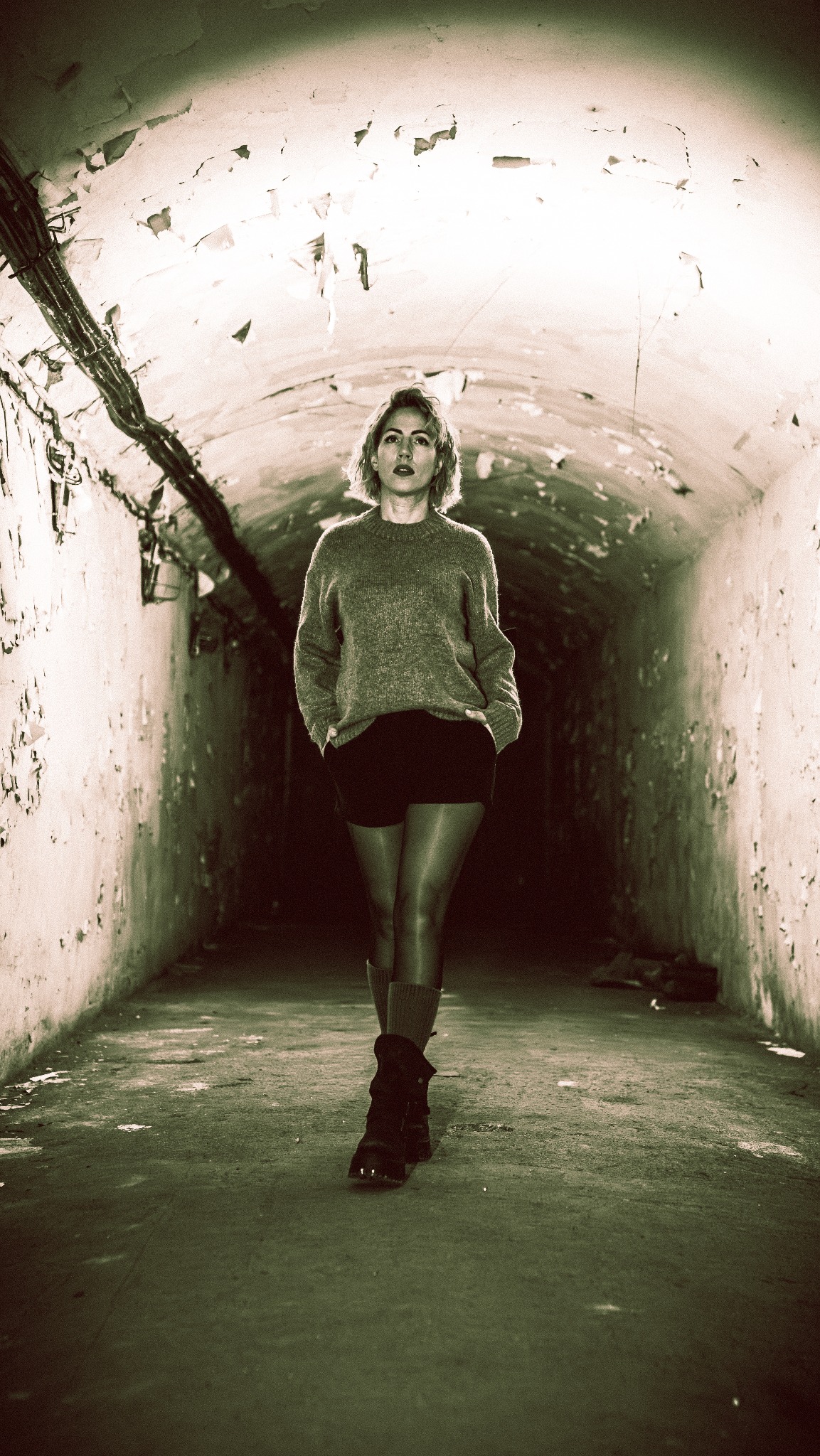 Kobieta w swetrze i szortach pozuje w tunelu z odpadającą farbą, oświetlonym od góry. Ujęcie centralne, podkreślające surowość otoczenia i kontrast z postacią.