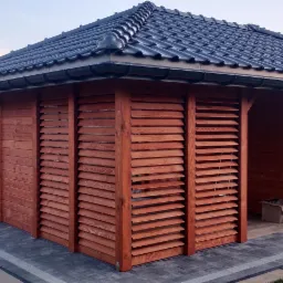 Drewniana altana z żaluzjowymi ścianami i czarnym dachem z dachówki ceramicznej, wykończona kostką brukową na tle budującego się domu.