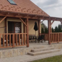 Drewniana konstrukcja zadaszenia tarasu przylegająca do domu z pomarańczową dachówką, widoczne meble ogrodowe i czarny parasol, obok zaparkowany samochód.