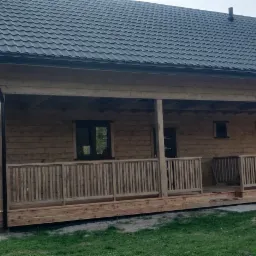 Gotowy dom z drewnianych bali z zadaszonym tarasem, widoczne ciemne okna i metalowy dach, otoczony zielenią.
