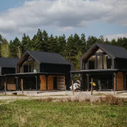 Czarne domy modułowe z płaskimi dachami w surowym stanie, otoczone zielenią i drzewami. Widoczne elementy konstrukcyjne i wykończeniowe.