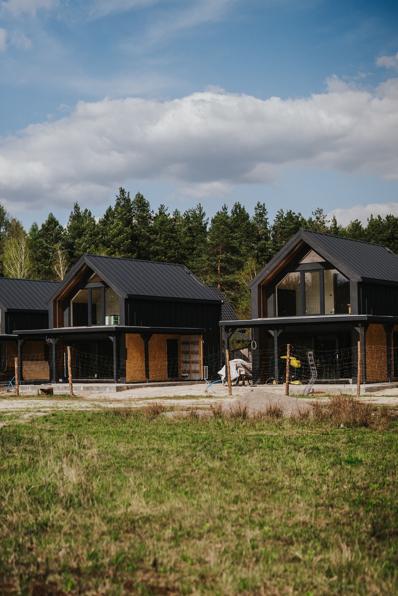 Czarne domy modułowe z płaskimi dachami w surowym stanie, otoczone zielenią i drzewami. Widoczne elementy konstrukcyjne i wykończeniowe.