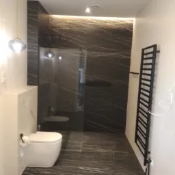 Nowoczesna łazienka z czarnymi płytkami imitującymi marmur na ścianach i podłodze, biała toaleta z zabudowaną spłuczką, czarny grzejnik drabinkowy i szklana kabina prysznicowa.