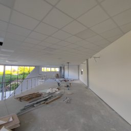 BUD-REM SYSTEM - Wnętrze w trakcie wykończenia: sufit podwieszany z płyt, gładkie ściany, widoczne narzędzia i materiały budowlane. Duże okna z widokiem na zieleń.