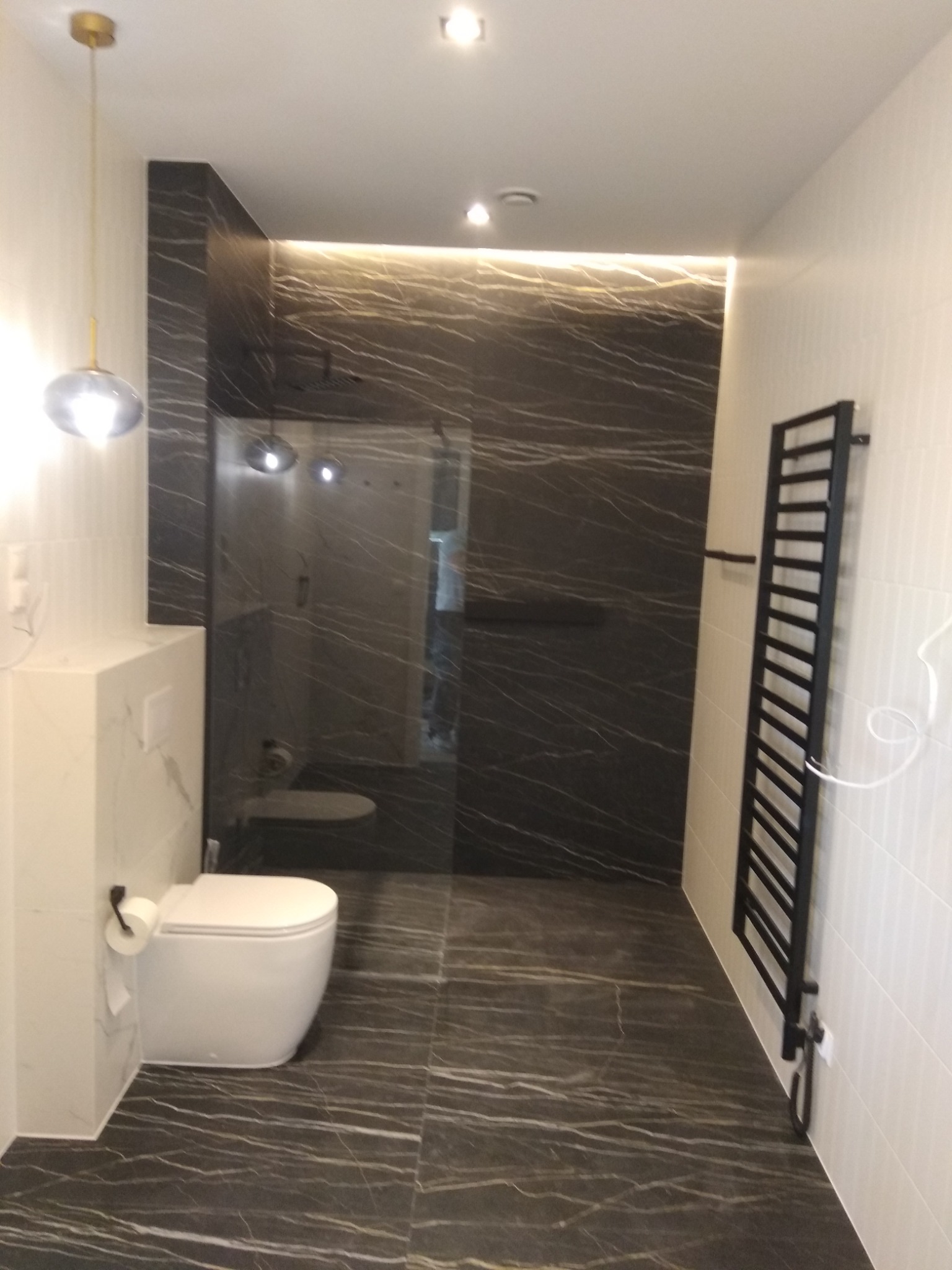 Nowoczesna łazienka z czarnymi płytkami imitującymi marmur na ścianach i podłodze, biała toaleta z zabudowaną spłuczką, czarny grzejnik drabinkowy i szklana kabina prysznicowa.