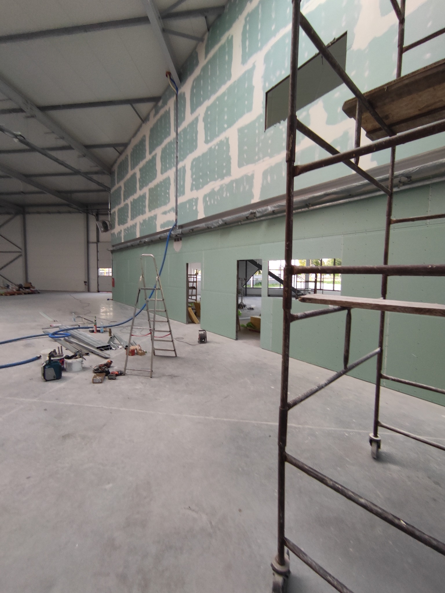 Montaż płyt G-K w hali: ściana z widocznymi łączeniami, rusztowanie, drabina i narzędzia na betonowej posadzce. Widoczne otwory drzwiowe i okienne w ścianie.