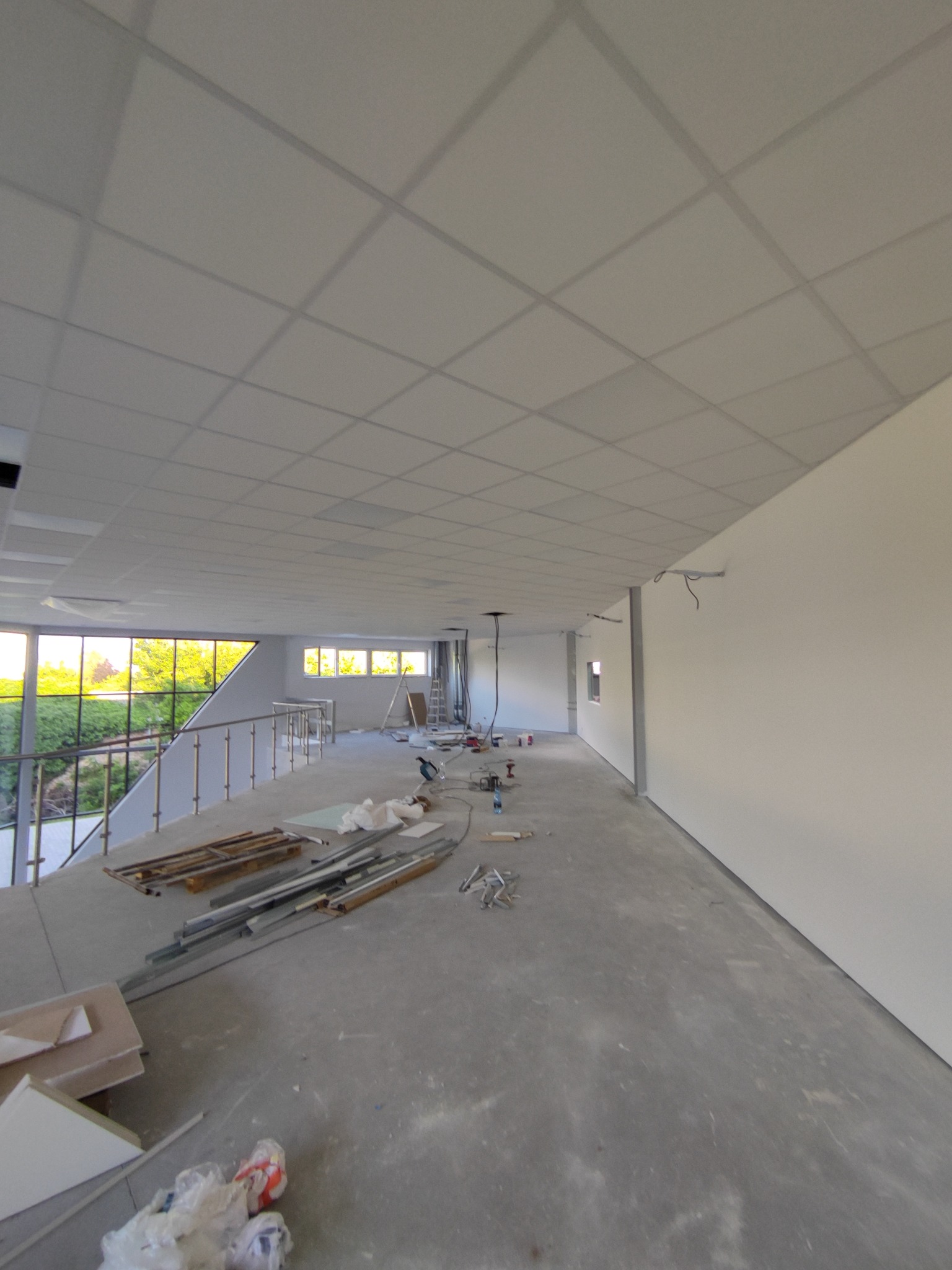 Wnętrze w trakcie wykończenia: sufit podwieszany z płyt, gładkie ściany, widoczne narzędzia i materiały budowlane. Duże okna z widokiem na zieleń.