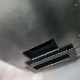 Metalowa obudowa wymiennika ciepła powietrze-powietrze w trakcie montażu, widoczne otwory wentylacyjne i śruby mocujące, z czerwonym statywem w tle.