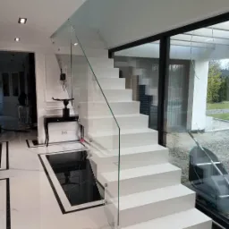 Nowoczesne, białe schody metalowe z szklaną balustradą w luksusowym wnętrzu, z widoczną czarną konsolą i marmurową podłogą.