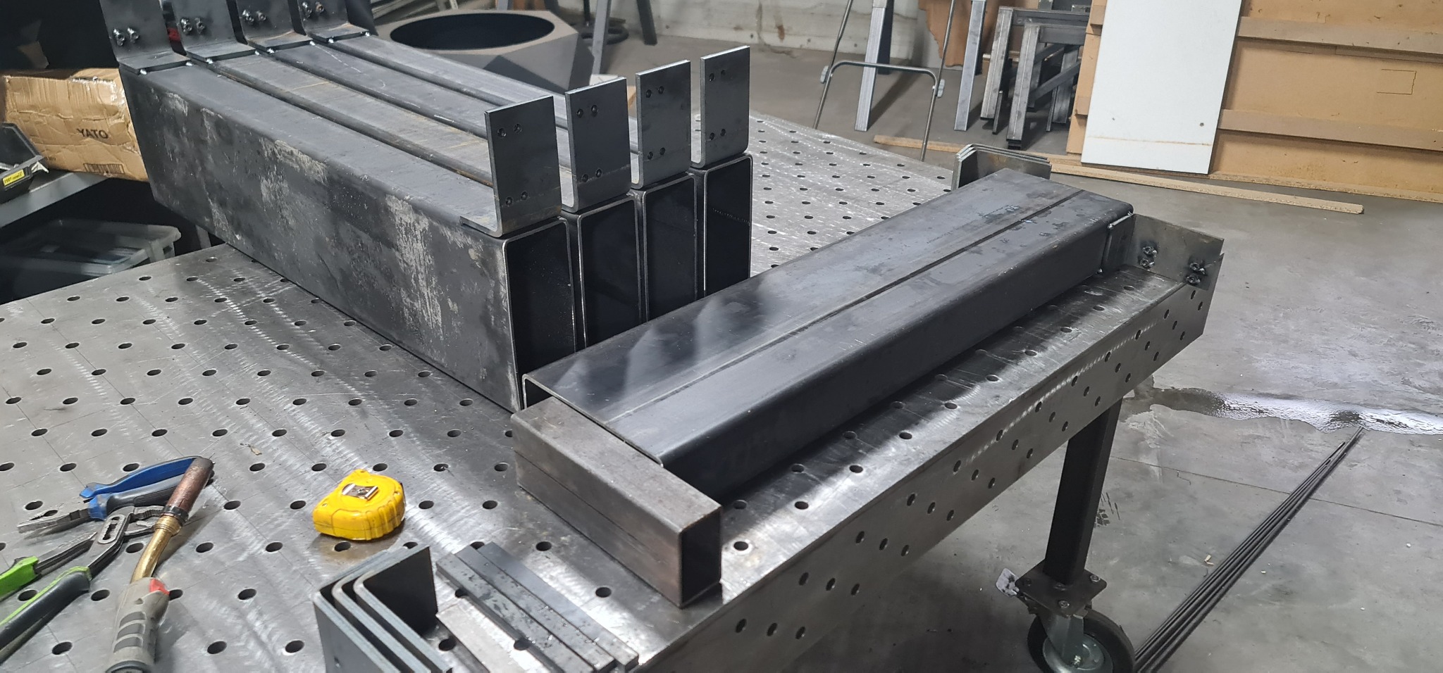 Przygotowane elementy konstrukcji z profili stalowych na perforowanym stole spawalniczym, widoczne narzędzia i miarka.