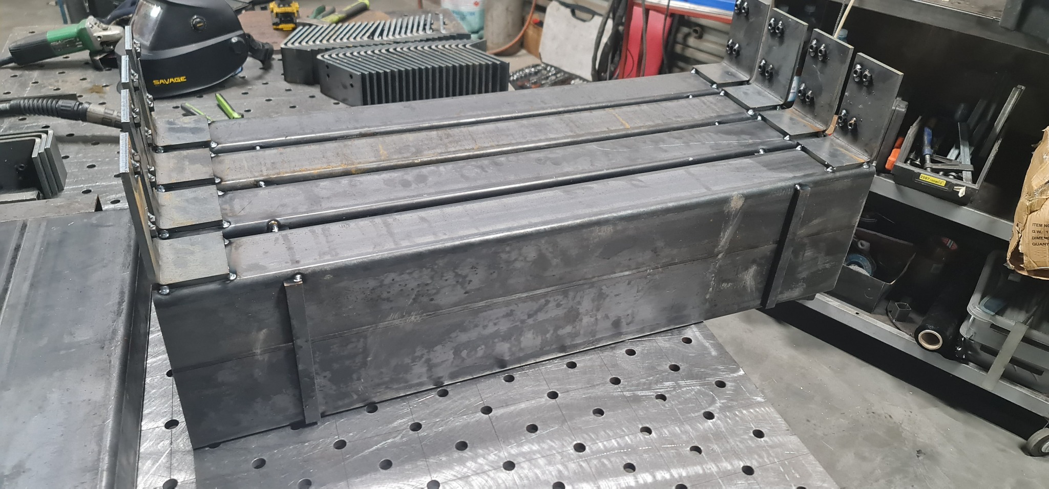 Surowe, spawane profile stalowe z przyspawanymi elementami mocującymi, ułożone na perforowanym stole spawalniczym, z widocznym wyposażeniem warsztatu w tle.