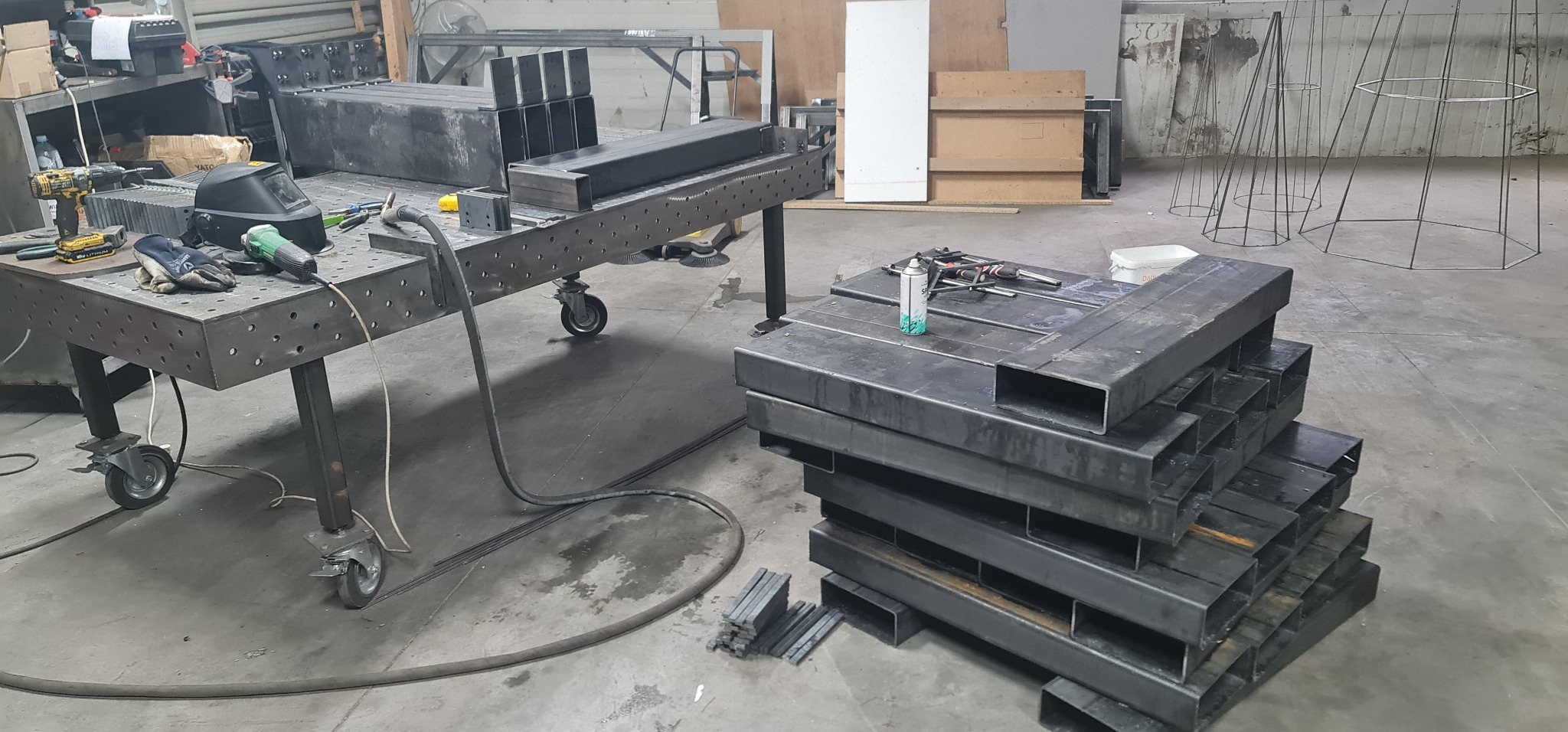 Warsztatowa scena z metalowymi profilami ułożonymi w stos, obok stołu spawalniczego z narzędziami i maską, oraz konstrukcjami metalowymi w tle.