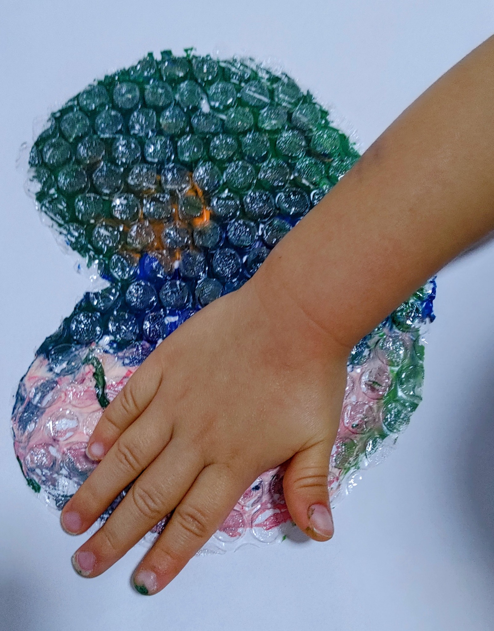 Dziecięca dłoń dotyka malowidła wykonanego na folii bąbelkowej, farby w kolorach zieleni, niebieskiego i różu tworzą abstrakcyjny wzór.