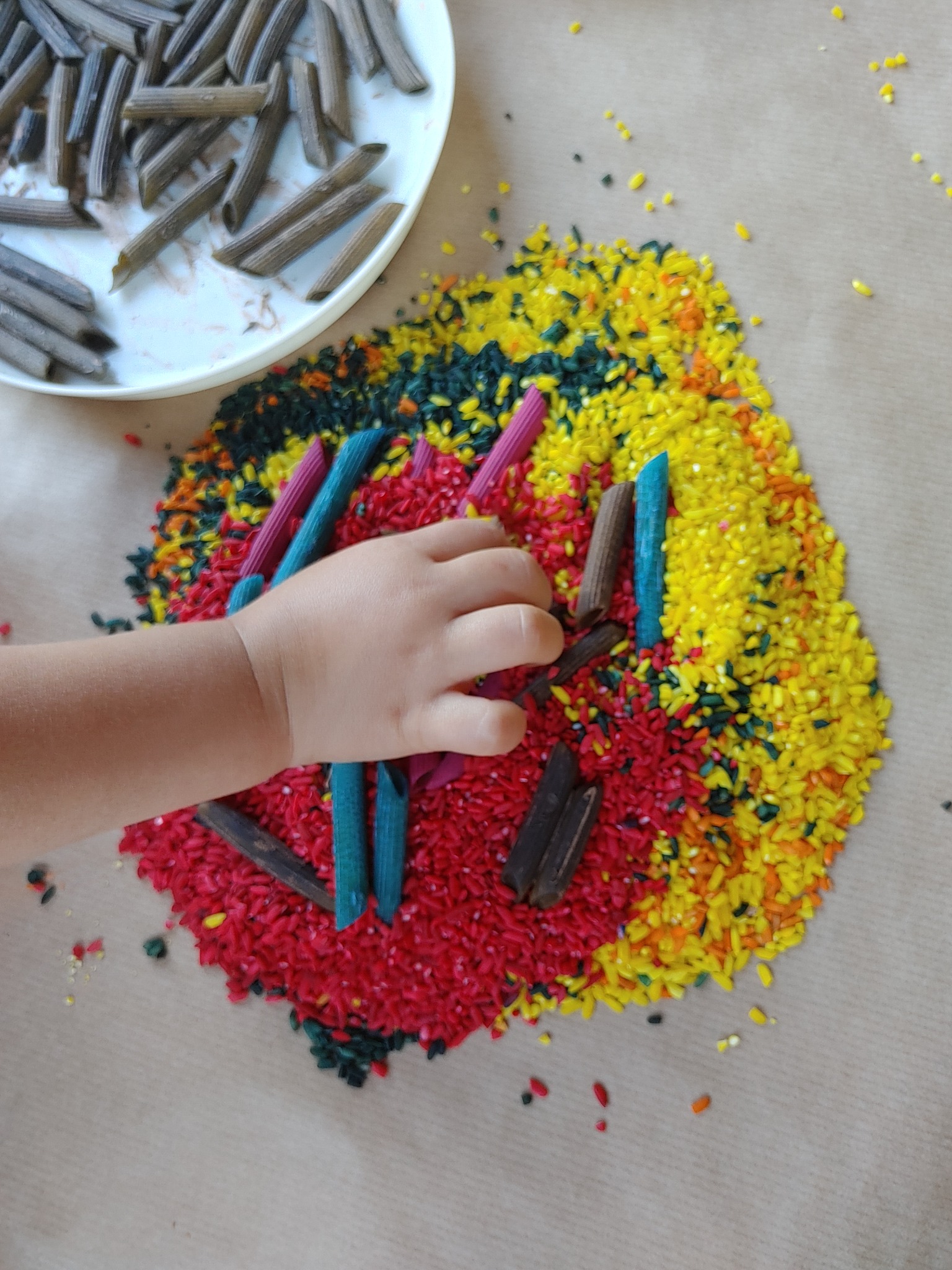Dziecięca dłoń układa kolorowe makarony penne na tle barwnego ryżu, tworząc wzór na brązowym papierze. Część makaronów leży na białym talerzu.