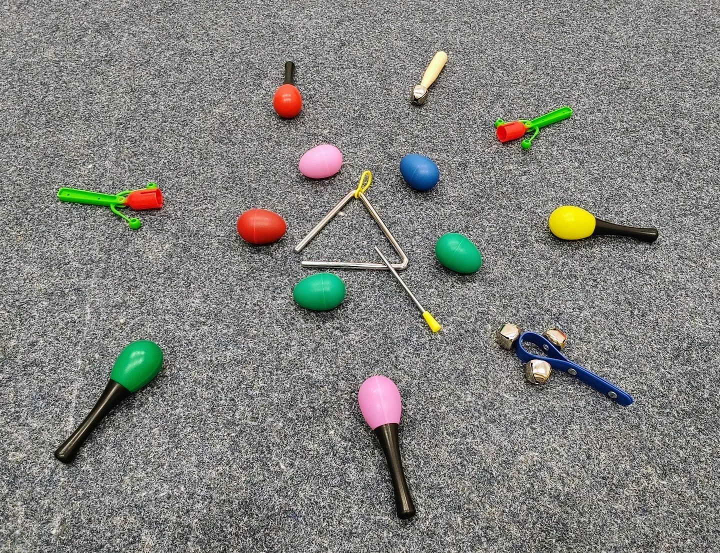 Różnorodne instrumenty perkusyjne, w tym marakasy w kształcie jajek, trójkąt, dzwonki i grzechotki, ułożone na szarym dywanie.