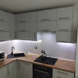 Nowoczesna kuchnia z białymi szafkami i podświetleniem LED pod szafkami wiszącymi, drewniany blat, czarny zlew i płyta grzewcza. Widoczne gniazdko elektryczne.