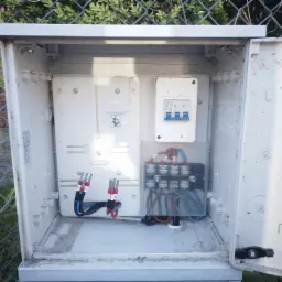 Otwarta, zewnętrzna skrzynka elektryczna z zabezpieczeniami, bezpiecznikami i przewodami. Widoczny fragment ogrodzenia z siatki i ceglany mur w tle.