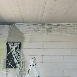 Bartosz Grzanka - Białe kable elektryczne ułożone równolegle pod sufitem surowego wnętrza z szarą ścianą z bloczków i aluminiową drabiną. Widoczna puszka instalacyjna.