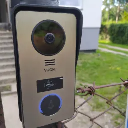 Zewnętrzny panel domofonowy VIRONE z kamerą i czytnikiem kart zbliżeniowych zamontowany na zardzewiałej metalowej siatce ogrodzeniowej na tle zieleni.