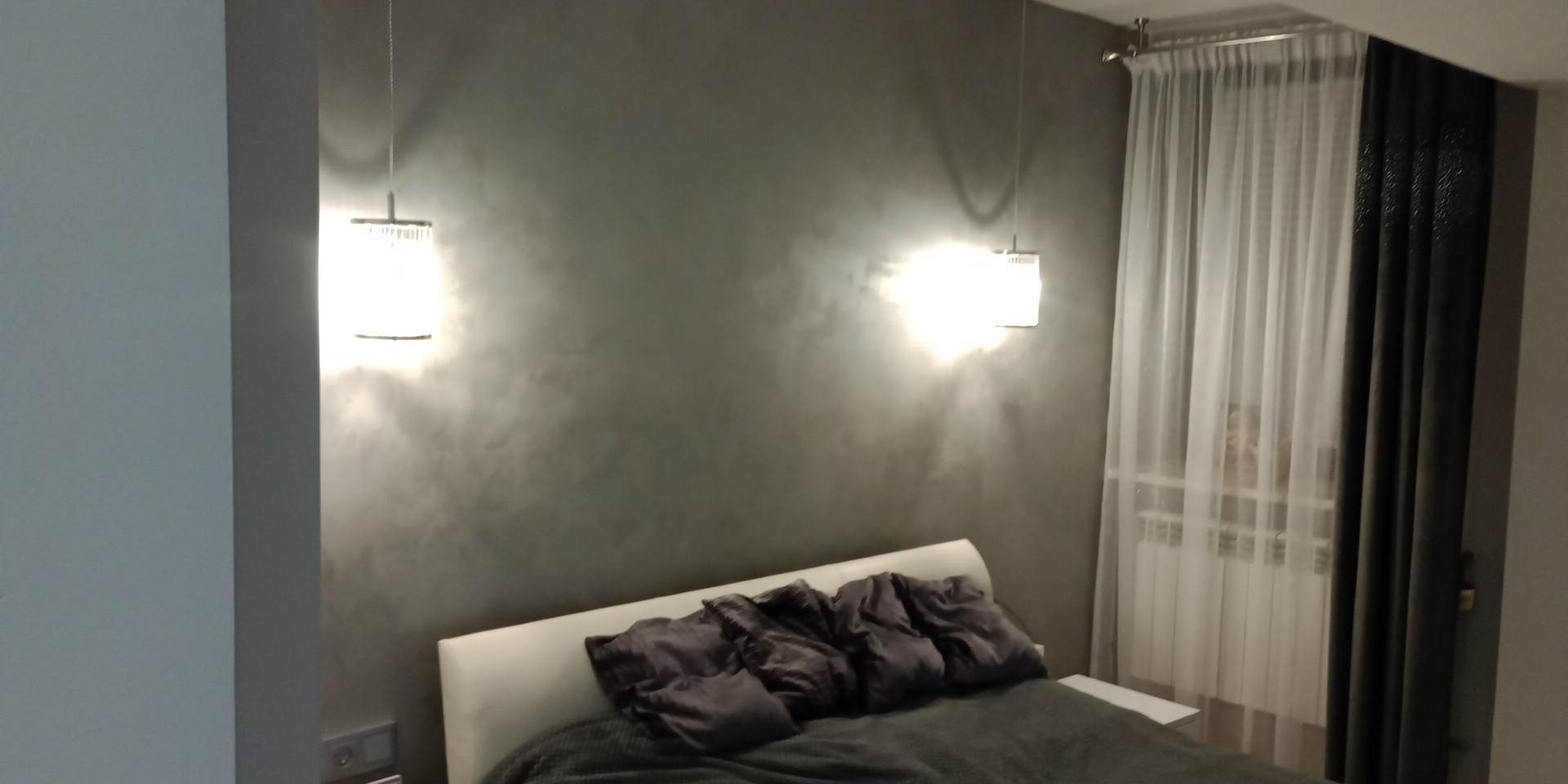 Sypialnia z szarą ścianą, nad łóżkiem wiszą dwie nowoczesne lampy z białymi abażurami. Okno zasłonięte firankami i zasłonami. Dyskretne oświetlenie tworzy przytulny nastrój.