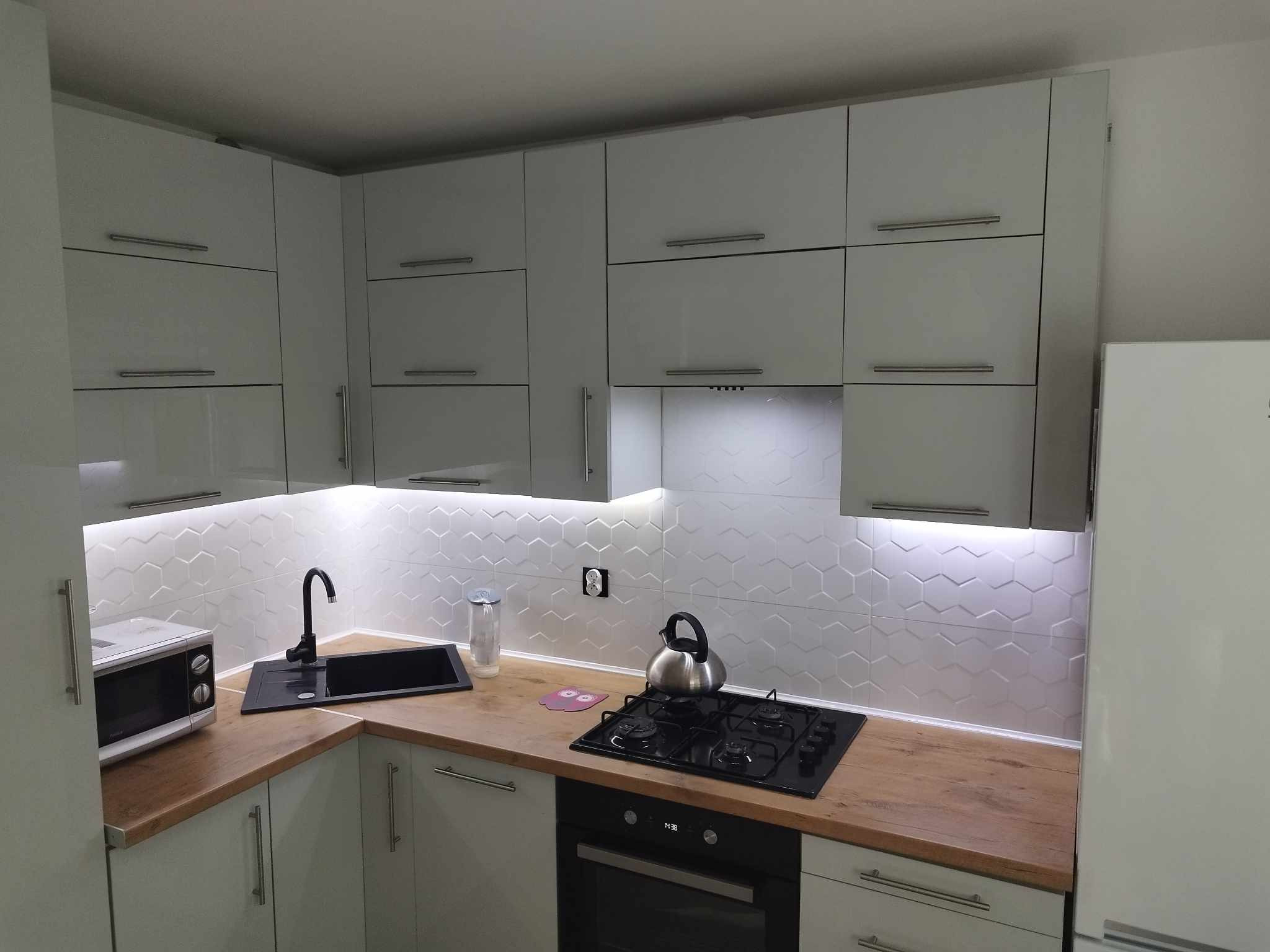 Nowoczesna kuchnia z białymi szafkami i podświetleniem LED pod szafkami wiszącymi, drewniany blat, czarny zlew i płyta grzewcza. Widoczne gniazdko elektryczne.