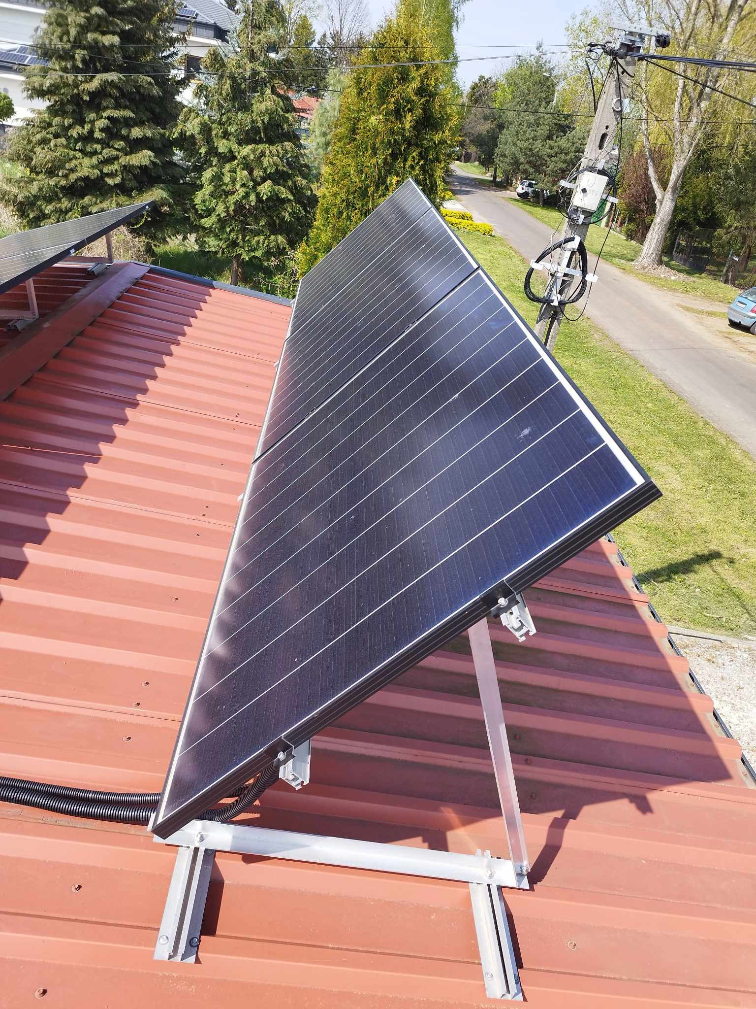 Panel słoneczny na metalowym dachu, zamontowany na aluminiowej konstrukcji. W tle widoczne drzewa i ulica. Instalacja fotowoltaiczna w słoneczny dzień.