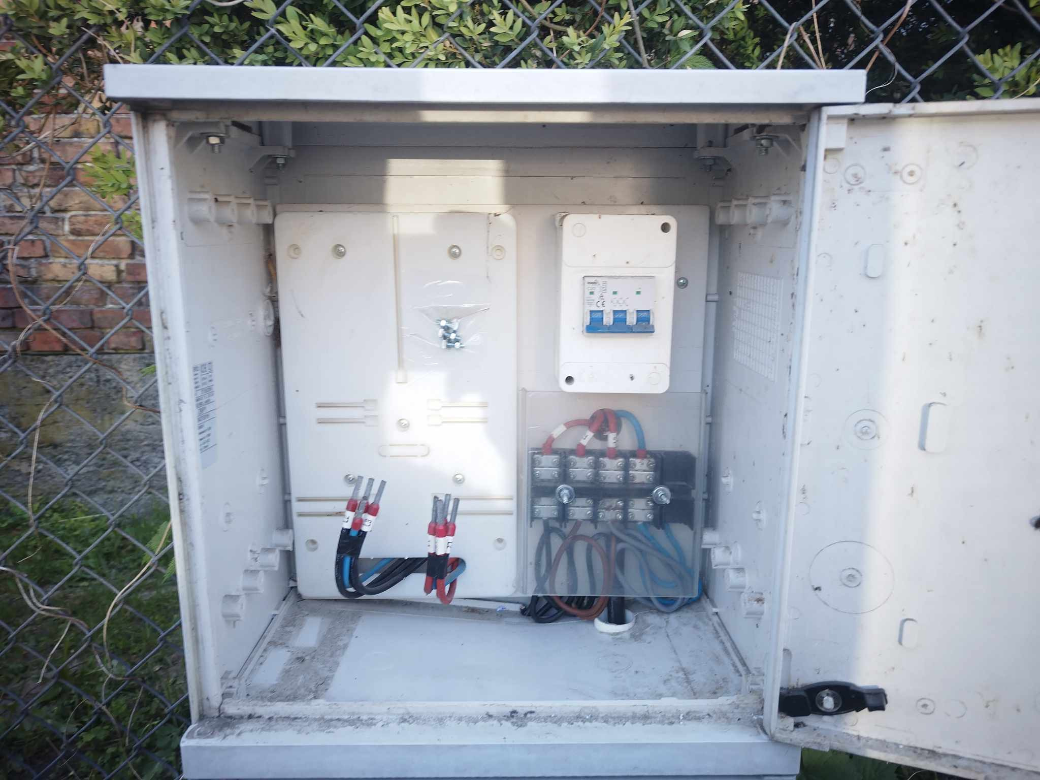Otwarta, zewnętrzna skrzynka elektryczna z zabezpieczeniami, bezpiecznikami i przewodami. Widoczny fragment ogrodzenia z siatki i ceglany mur w tle.