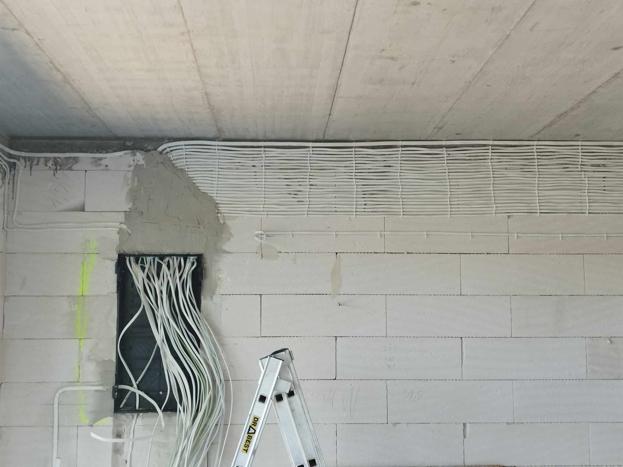 Białe kable elektryczne ułożone równolegle pod sufitem surowego wnętrza z szarą ścianą z bloczków i aluminiową drabiną. Widoczna puszka instalacyjna.