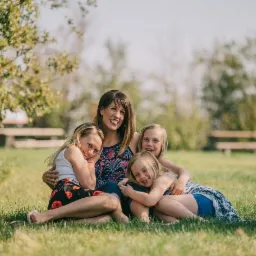 Portret uśmiechniętej kobiety z trzema córkami siedzącymi na trawie w słoneczny dzień, widoczne drzewa i ławka w tle.
