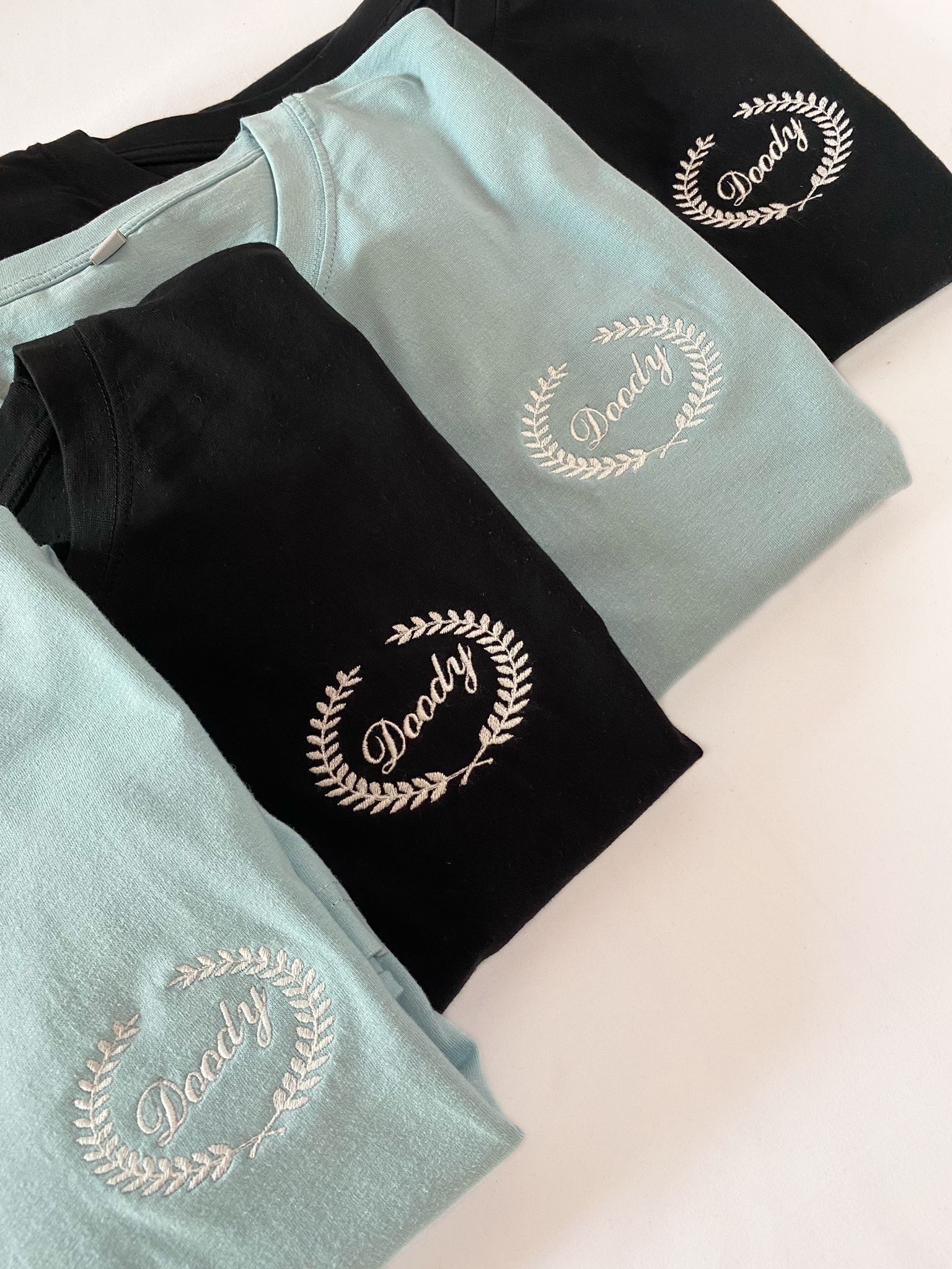 Stos złożonych t-shirtów w kolorach czarnym i błękitnym, każdy z wyhaftowanym białym napisem 'Doody' otoczonym wieńcem laurowym po lewej stronie klatki piersiowej.